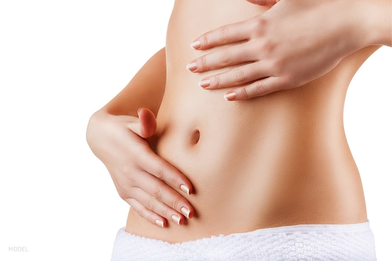 Woman touching her slim, bare abdomen with both of her hands.